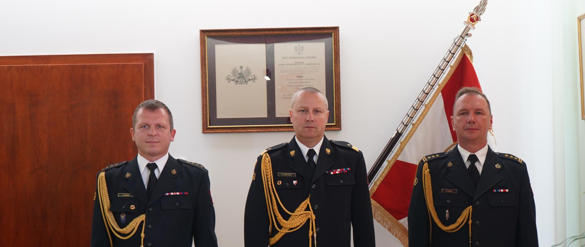 trzech strażaków w ciemnych mundurach ze sznurem stoi obok siebie, w tle sztandar 