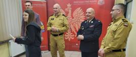 Strażacy organizatorzy turnieju stoją podczas wręczania dyplomów okolicznościowych na tle ścianki PSP. Jedna z uczestniczek odbiera dyplom.