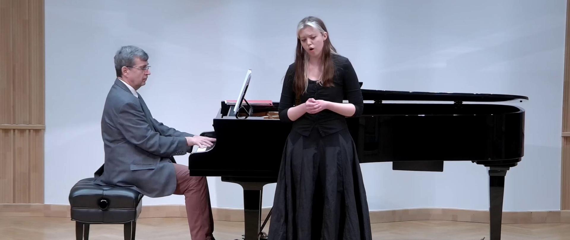 Zdjęcie. Na estradzie występuje ubrana w czarny strój śpiewaczka z towarzyszeniem pianisty, który gra na dużym czarnym fortepianie.