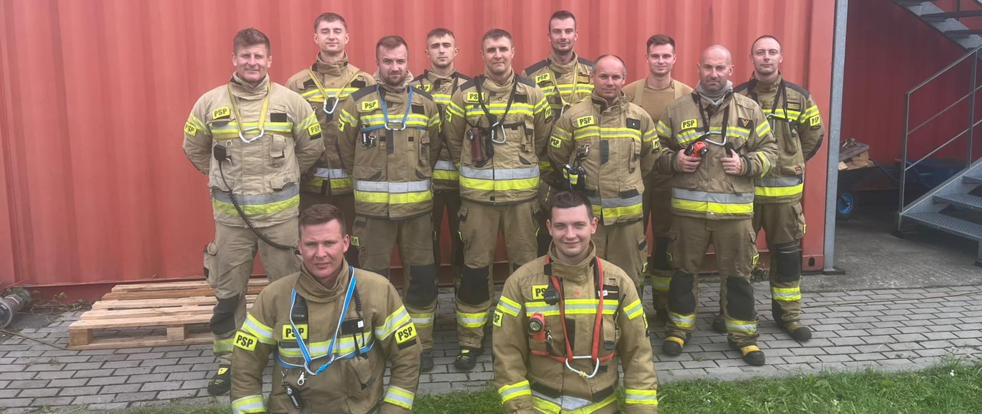 Dwunastu strażaków PSP pozuje do zdjęcia przed czerwonym kontenerem szkoleniowym. Dziesięciu stoi w rzędzie, dwóch klęczy z przodu. Wszyscy mają ubrania specjalne z odblaskami. Przed strażakami leżą żółte torby i sprzęt wykorzystywany podczas ćwiczeń RIT. W tle widać kontenery, schody i elementy infrastruktury szkoleniowej. Zdjęcie dokumentuje udział funkcjonariuszy w warsztatach z zakresu organizacji i działania rot asekuracyjnych.