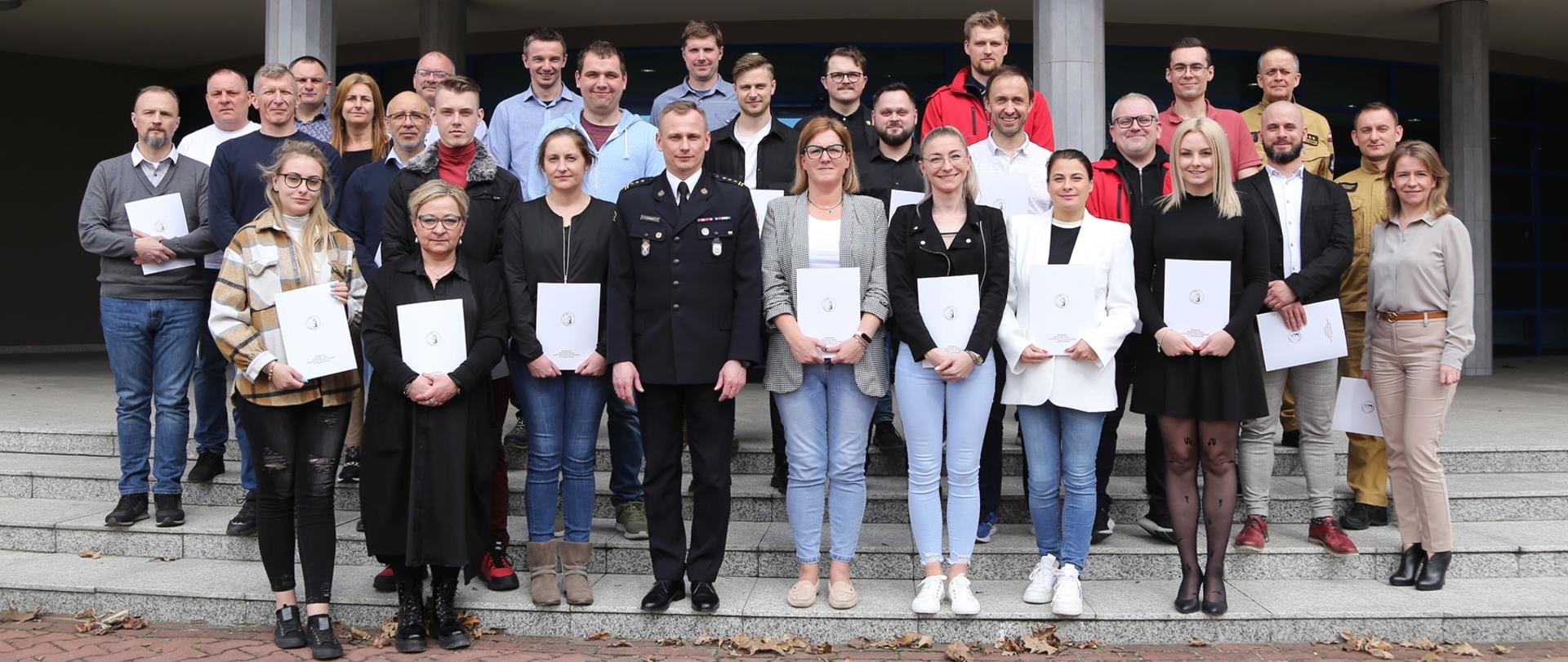 Grupowe zdjęcie uczestników szkolenia (26 osób) w towarzystwie Zastępcy Komendanta Centralnej Szkoły PSP, kobiety oraz dwóch funkcjonariuszy CS PSP