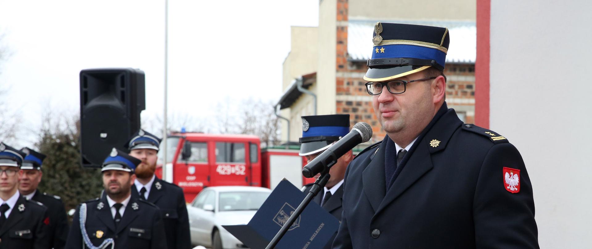 strażak mężczyzna w rogatywce i w płaszczu stoi przed mikrofonem , za nim inny strażak w rogatywce trzyma w ręku niebieską teczkę, w tle grupa strażaków w szeregu oraz samochód strażacki