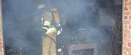 Pożar budynku mieszkalnego w Wydawach. Zadymione wnętrze pomieszczenia, w nim strażak prowadzący działania. Ma założony aparat oddechowy.