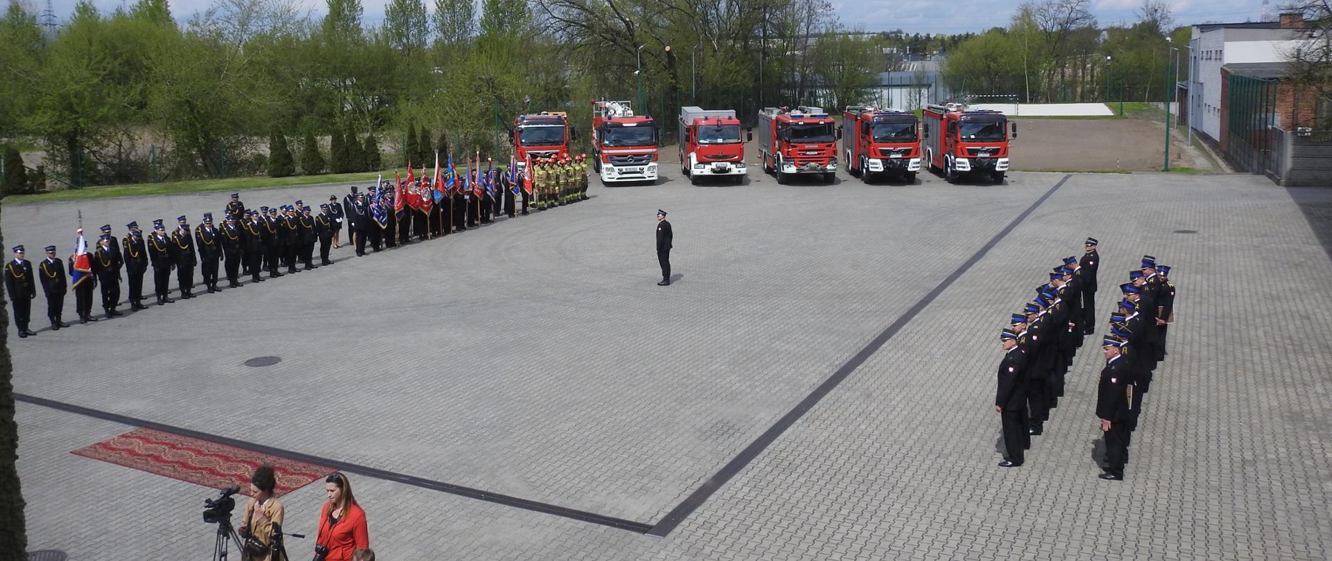 Zdjęcie zrobione z lotu ptaka. Duży plac. Po lewej stronie ustawieni strażacy i poczty sztandarowe. Po prawej stronie strażacy w dwuszeregu. Na wprost samochody strażackie. Na dole zdjęcia ludzie z aparatami i kamerami. W tle zielone drzewa.