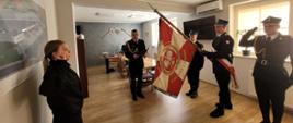 Zbiórka strażaków w jasnym pomieszczeniu. Po prawej stronie widoczny biało - czerwony sztandar komendy i 3 strażaków z pocztu sztandarowego. Po lewej kobieta w mundurze koszarowym składa ślubowanie. W środku komendant w mundurze wyjściowym odczytuje rotę ślubowania z granatowej teczki. W tle wyposażenie gabinetu.