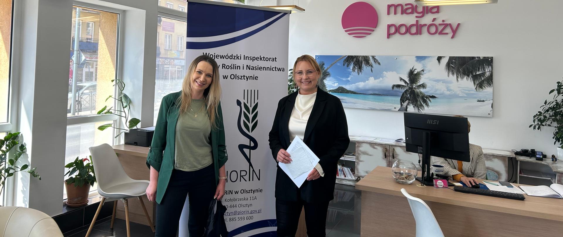 dwie kobiety stoją w biurze po obu stronach banera z napisem i logo WIORiN w Olsztynie, w tle logo firmy: Magia podróży