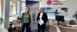 dwie kobiety stoją w biurze po obu stronach banera z napisem i logo WIORiN w Olsztynie, w tle logo firmy: Magia podróży