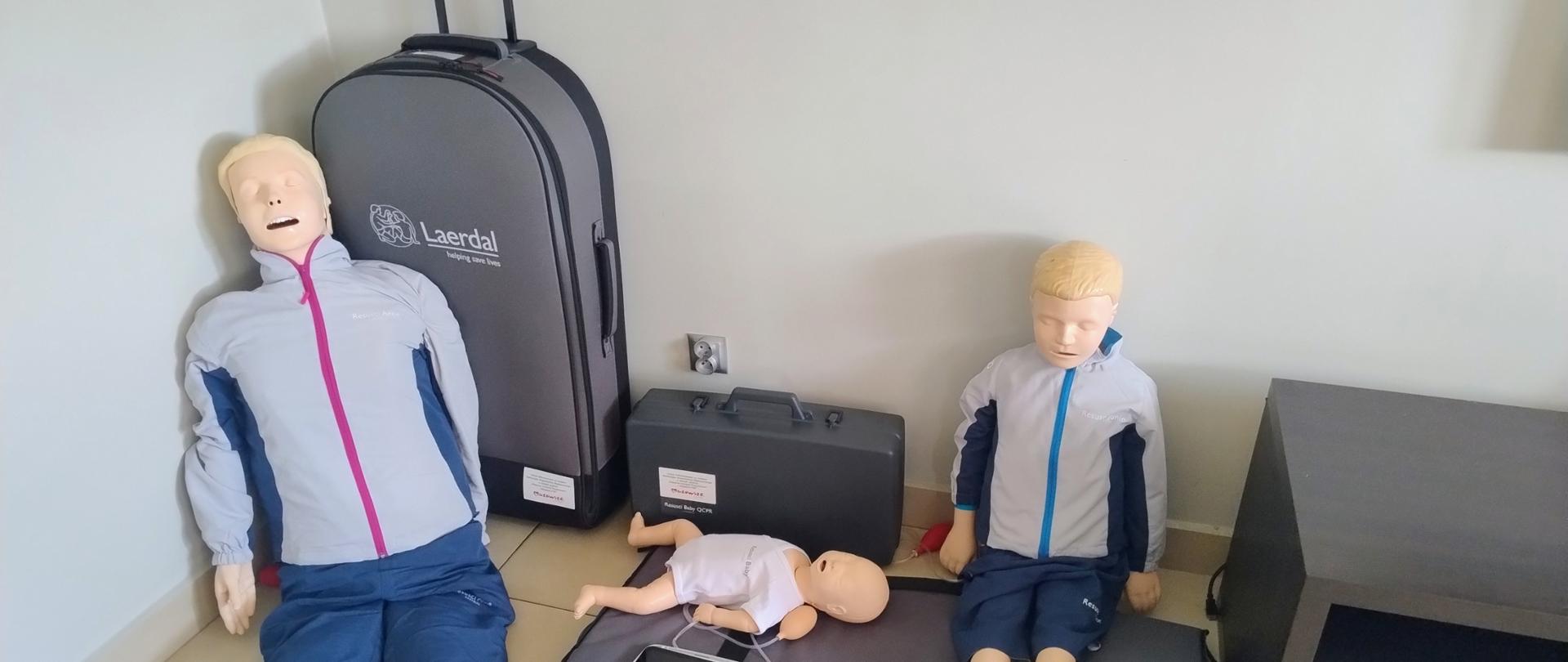 Zestaw fantomów szkoleniowych firmy Laerdal ustawiony przy ścianie. Na zdjęciu widoczny dorosły, dziecięcy i niemowlęcy fantom do nauki resuscytacji krążeniowo-oddechowej. Obok znajdują się torby transportowe oraz sprzęt dodatkowy.