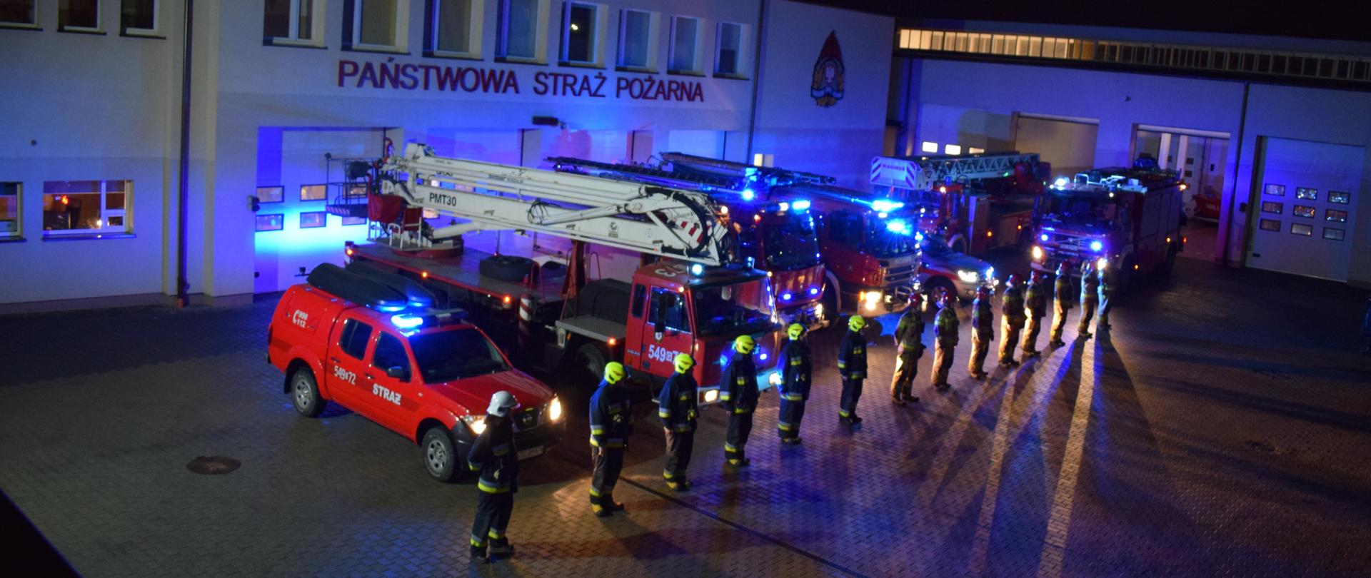 Plac JRG Tczew. Wieczór. Ustawione w szeregu pojazdy JRG Tczew i OSP Tczew z włączonymi światłami błyskowymi. Przed samochodami stoją strażacy w umundurowaniu specjalnym oddając hołd poległym strażakom z Czernikowa.