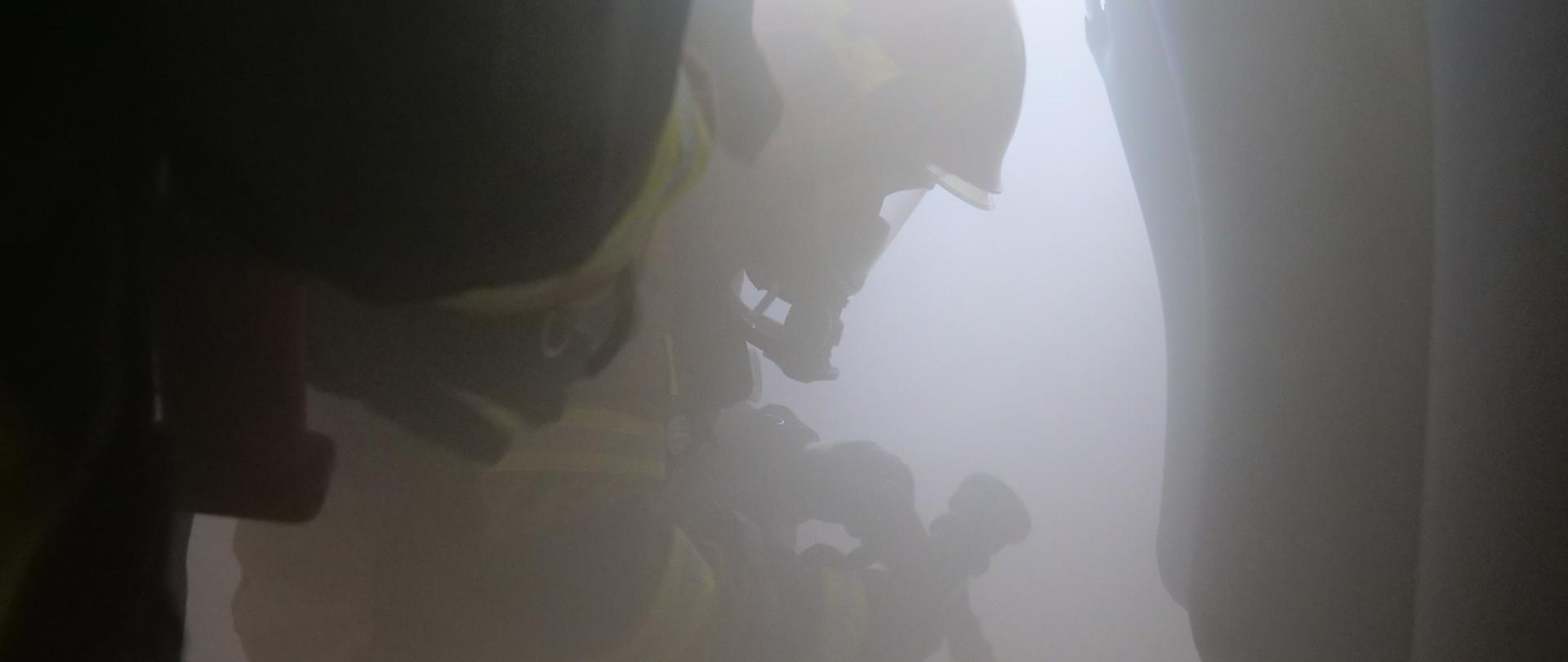 Kompletnie umundurowany strażak przyklęka na kolano trzymając prądownicę, wokół silne zadymienie.