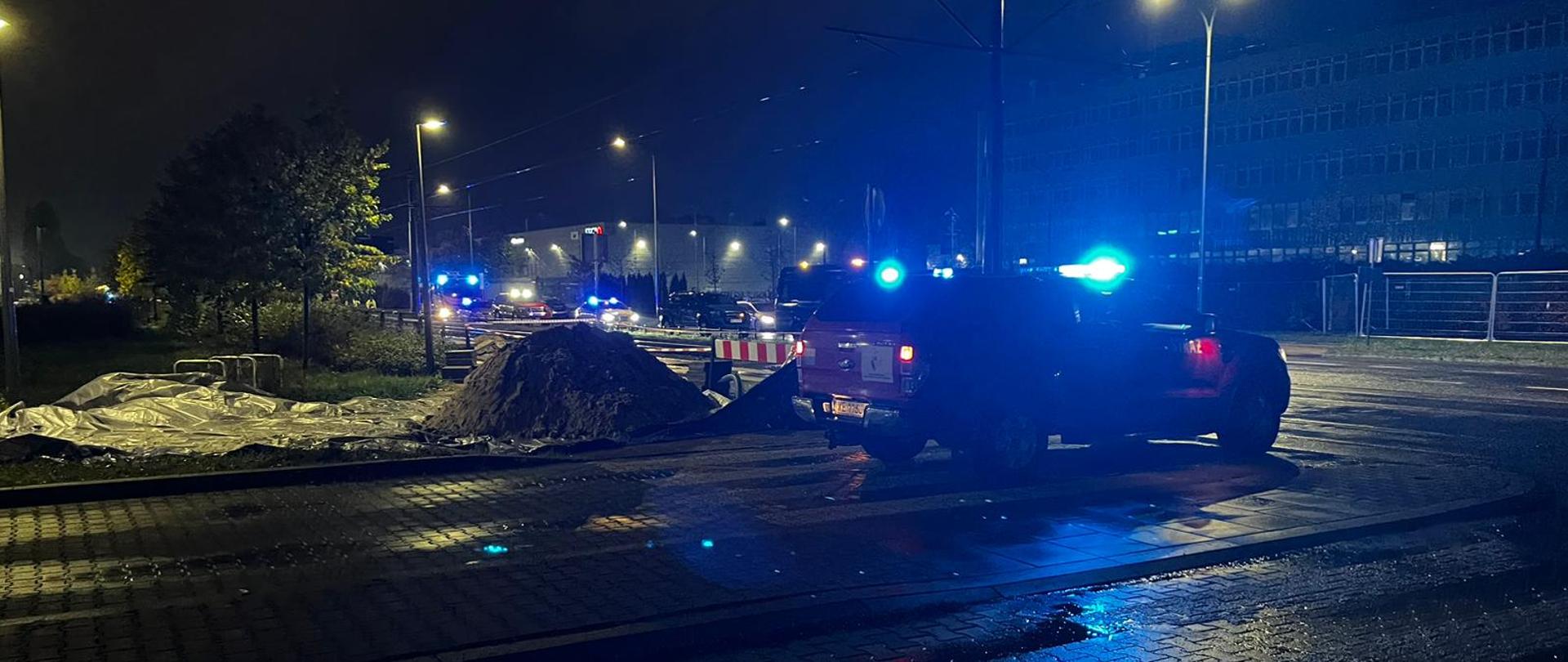 Skrzyżowanie. Na pierwszym planie widoczny samochód strażacki, obok na poboczu hałda ziemi z wykopu. Zdjęcie wykonane w porze nocnej.
