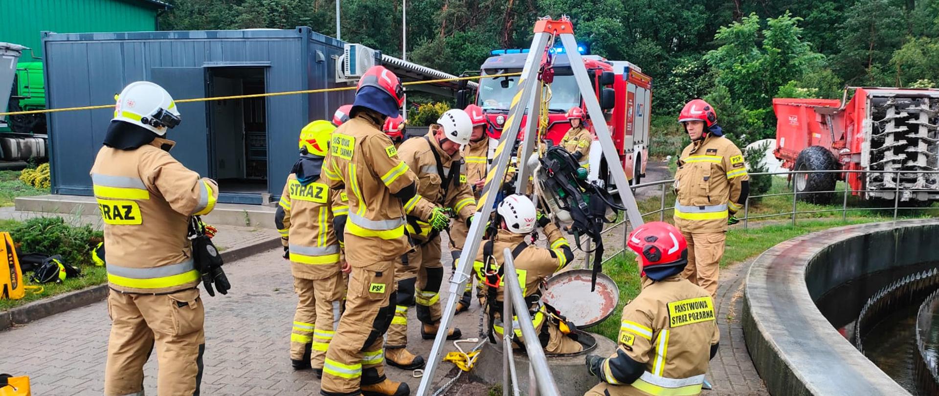 Ćwiczenia obiektowe na terenie oczyszczalni ścieków w Brzezinach, strażacy prowadzą działania aplikacyjne polegające na ewakuacji osoby poszkodowanej uwiezionej w studzience technologicznej, rozstawili trójnóg i przy jego pomocy jeden ze strażaków zjeżdża do studzienki w celu ewakuacji poszkodowanego, wokół trójnoga asekurują inni strażacy, w oddali widać samochód pożarniczy i infrastrukturę oczyszczalni ścieków.