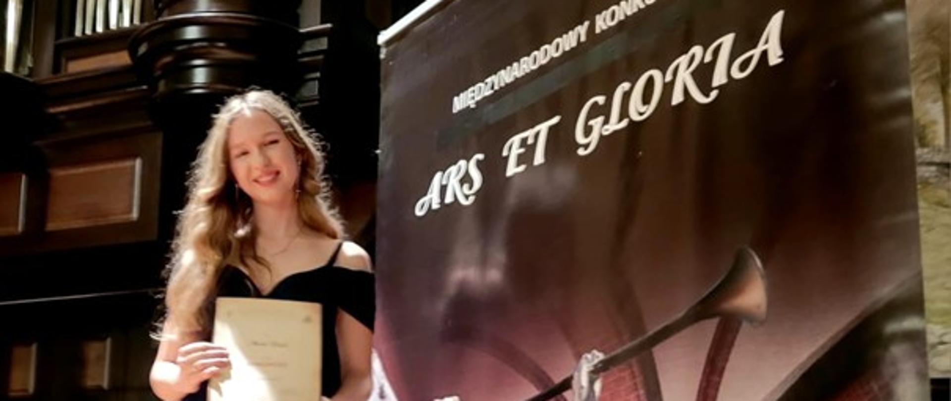 Amelia Szmidt - Ars Et Gloria 2025. Uczennica pozuje do zdjęcia przy plakacie konkursu.