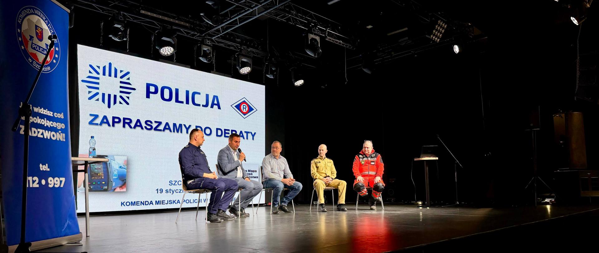 Obraz przedstawia panel dyskusyjny na scenie. Pięciu mężczyzn siedzi na krzesłach przed dużym ekranem, na którym widnieje logo i napis: „POLICJA ZAPRASZAMY DO DEBATY”. Na lewym banerze widoczny jest numer alarmowy 12 997. Uczestnicy panelu ubrani są w różne stroje, w tym mundur policyjny, cywilne ubrania, jasny mundur i czerwony kombinezon ratowniczy. Tło sceny jest czarne, a oświetlenie profesjonalne.