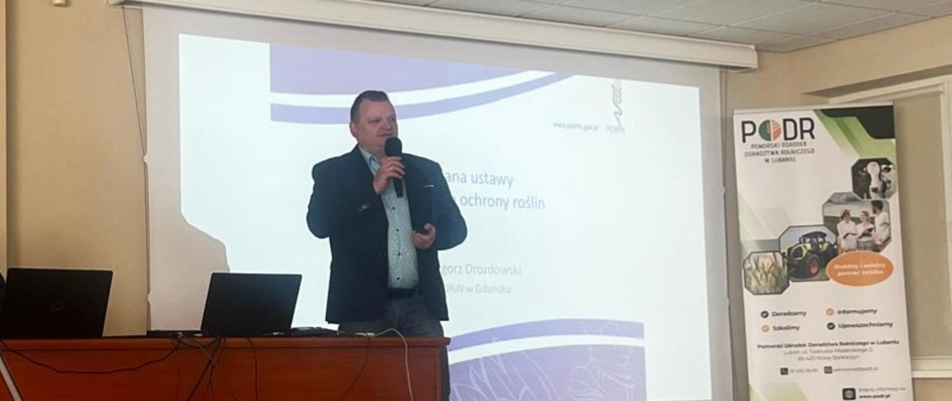 Mężczyzna w garniturze na scenie z mikrofonem, za nim na ścianie wyświetlona jest prezentacja dotycząca ustawy o środkach ochrony roślin, obok stoi biurko, na którym stoi otwarty laptop
