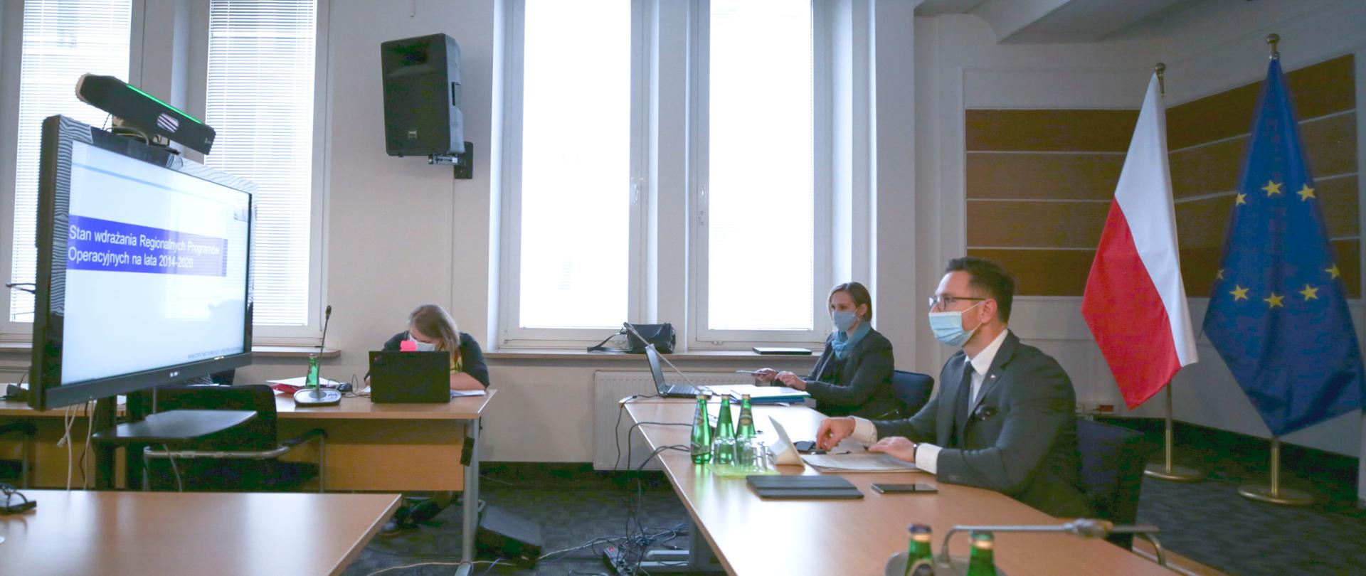 W sali konferencyjnej przy stołach siedzą trzy osoby. Pierwszy z prawej - wiceminister Waldemar Buda patrzy na duży monitor. Za nim dwie flagi polska i unijna.