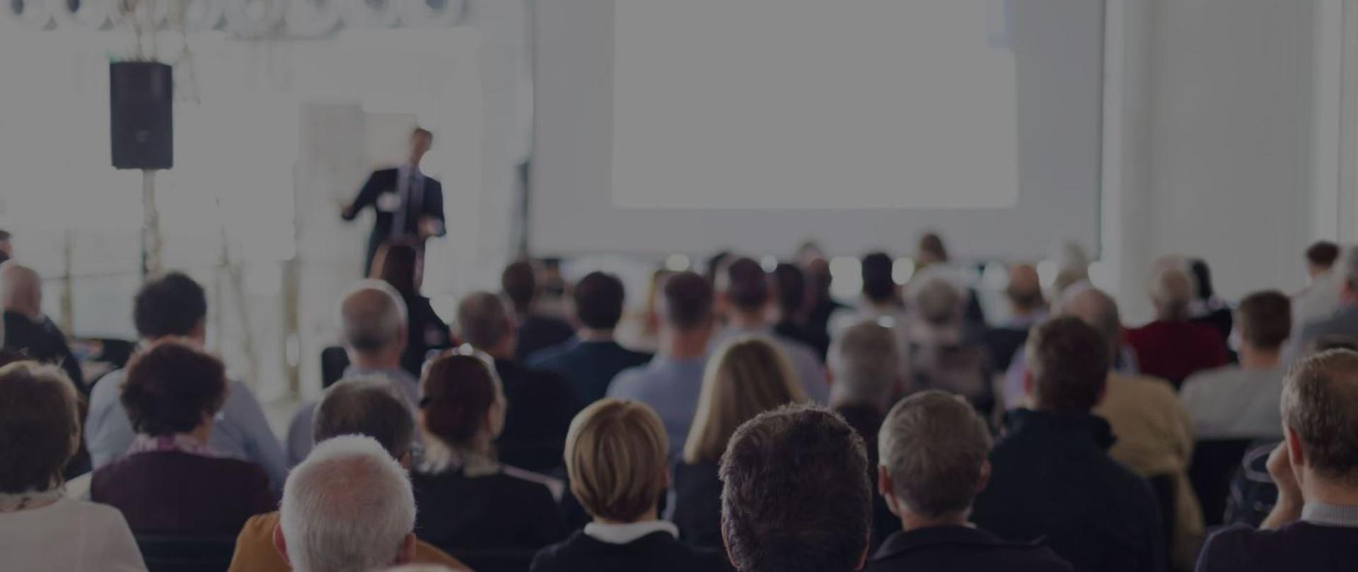 zdjęcie symbolizujące spotkanie - przed tablicą prelegent na którego patrzą siedzący na sali ludzie