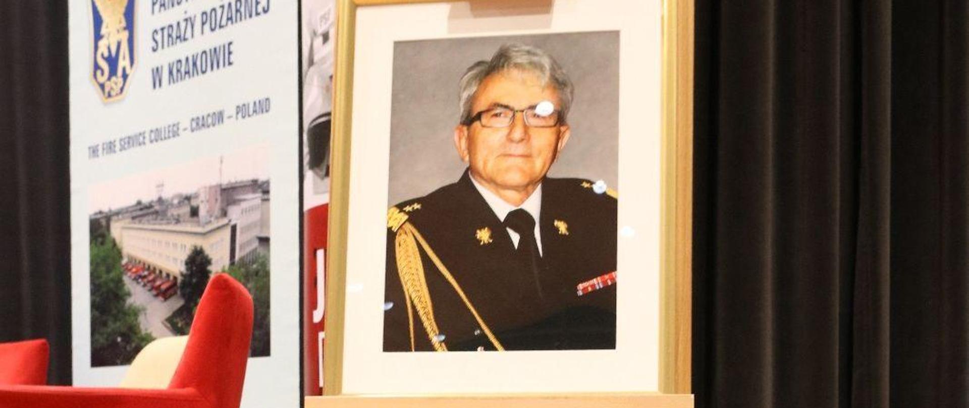 Zdjęcie gen. brygadiera Feliksa Deli w ramkach na sztaludze stoi na scenie. U podnóża sztalugi wiązanka biało czerwonych kwiatów.