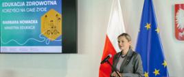 Kobieta w szarym garniturze i z blond włosami spiętymi w kok stoi za mównicą i przemawia do mikrofonu. W tle, po jej prawej stronie, znajdują się flagi Polski i Unii Europejskiej oraz godło Polski. Po lewej stronie widać duży ekran, na którym wyświetlany jest graficzny baner z napisem „Edukacja Zdrowotna. Korzyści na całe życie. Barbara Nowacka. Ministra Edukacji”. Na pierwszym planie widoczne są fragmenty głów publiczności.