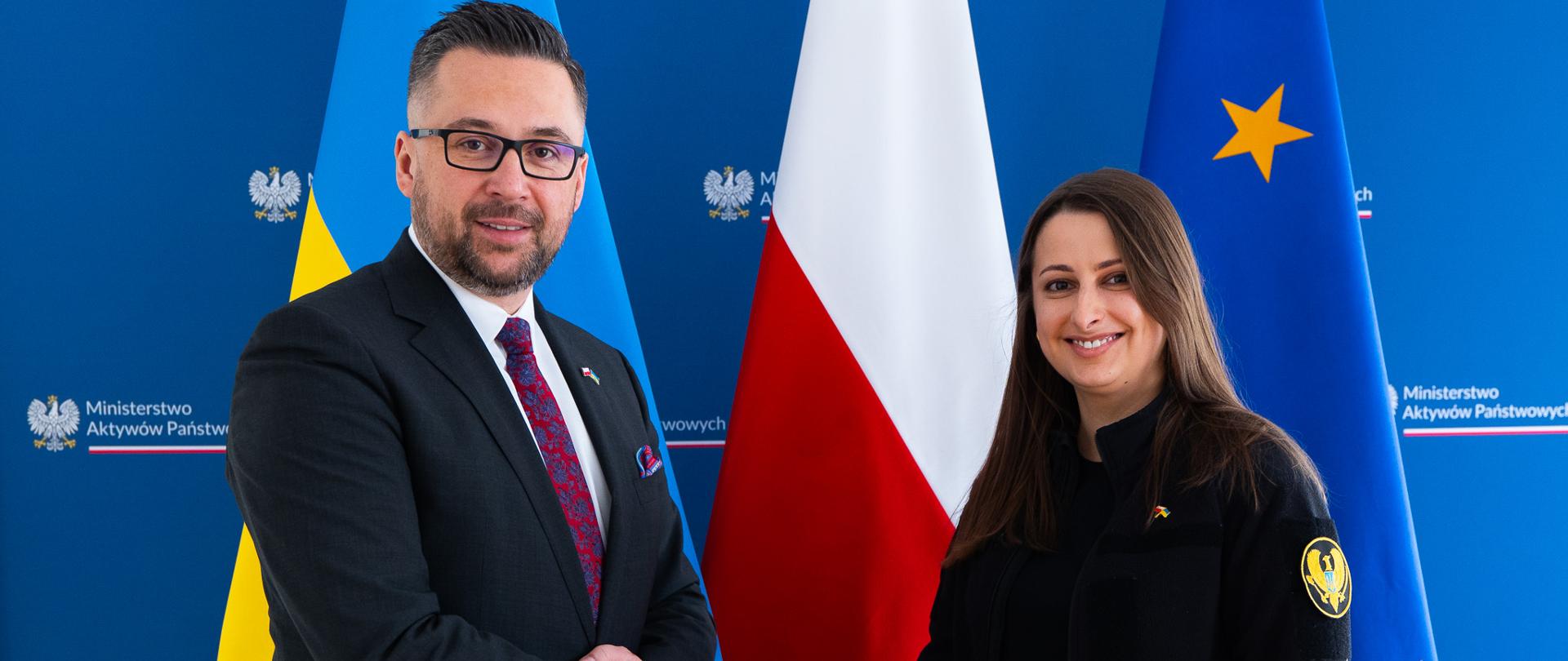 Wiceminister Kulasek stoi po lewej stronie zdjęcia i podaje rękę wiceminister Annie Gvozdiar, która stoi po prawej. W tle - tekstylna ścianka z logotypami MAP oraz trzy flagi. Od lewej to kolejno: Flaga Ukrainy, flaga Polski i flaga Unii Europejskiej.