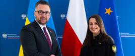 Wiceminister Kulasek stoi po lewej stronie zdjęcia i podaje rękę wiceminister Annie Gvozdiar, która stoi po prawej. W tle - tekstylna ścianka z logotypami MAP oraz trzy flagi. Od lewej to kolejno: Flaga Ukrainy, flaga Polski i flaga Unii Europejskiej.
