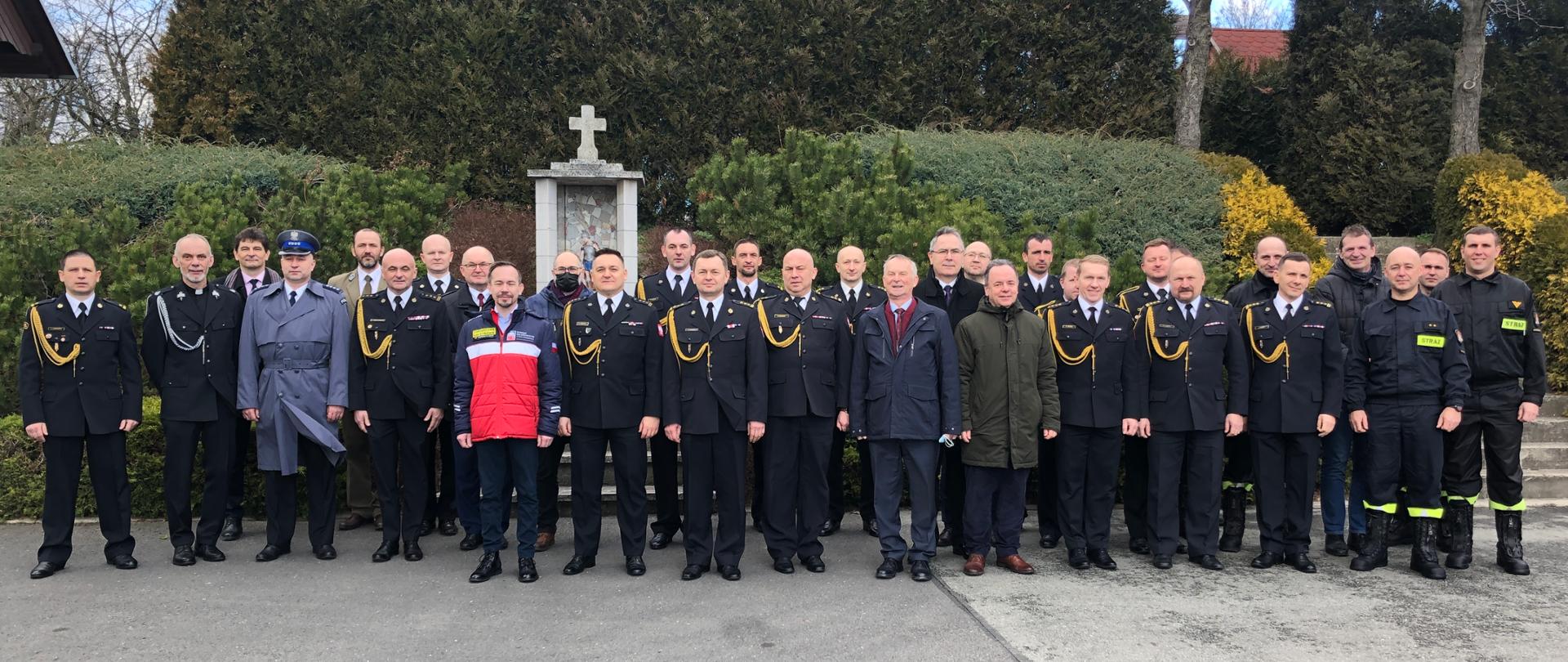 Zbiorowe zdjęcie uczestników uroczystej zbiórki z okazji zdania i przejęcia obowiązków komendanta powiatowego PSP w Krapkowicach. Na tle roślinności ustawieni uczestnicy uroczystości. W centrum - zdający obowiązki st. bryg. Krzysztof Kuniec, a także Opolski komendant Wojewódzki - st.bryg. Krzysztof Kędryk i pełniący obowiązki bryg. Rafał Bisowski oraz starosta krapkowicki - Maciej Sonik. 