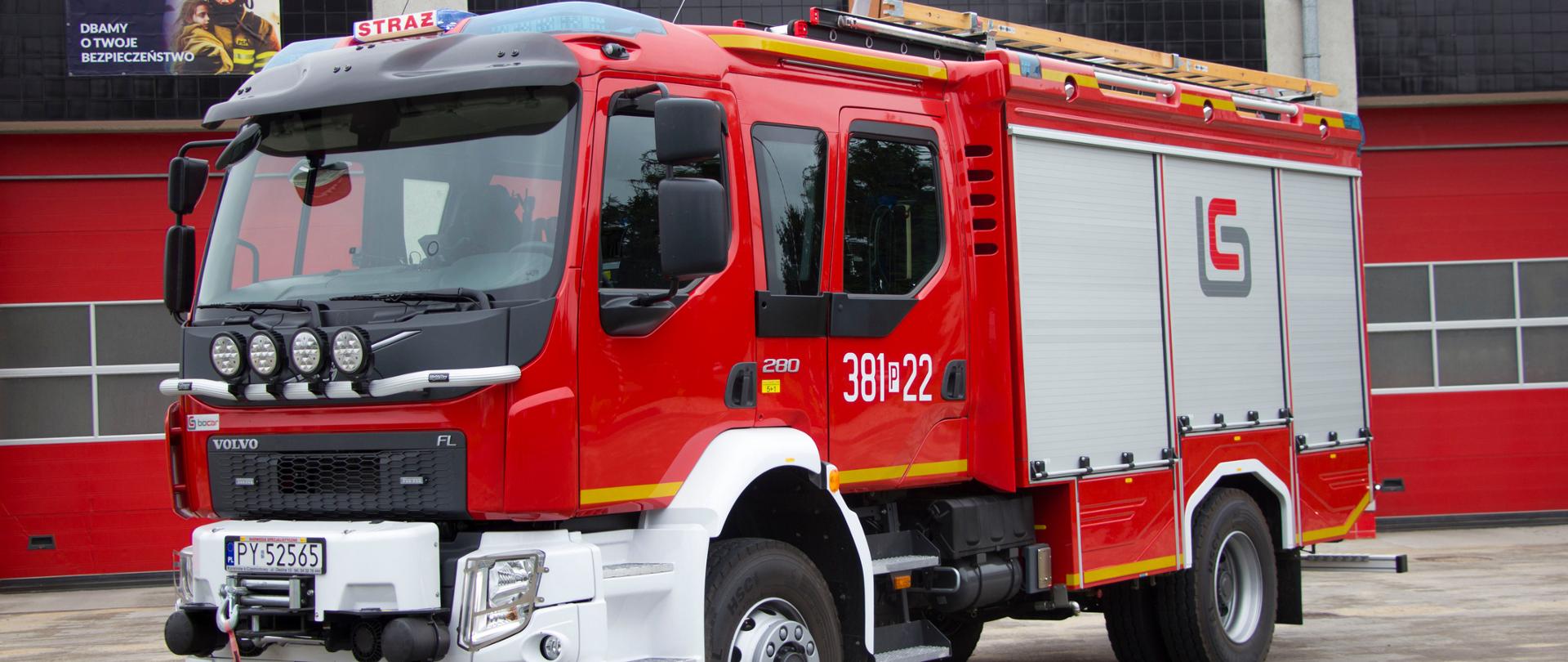 Duży samochód strażacki stoi przodem lekko z boku na placu na tle garaży komendy.