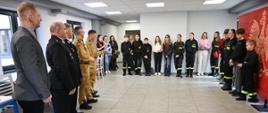 Zdjęcie przedstawia grupę osób stojących w półokręgu w jasnym pomieszczeniu. Po lewej stronie widoczni są dorośli mężczyźni, w tym kilku w mundurach strażackich, a po prawej młodzież i dzieci – część z nich ubrana w czarne stroje z odblaskowymi elementami, przypominające mundury strażackie. W tle znajduje się czerwony baner z białym orłem
