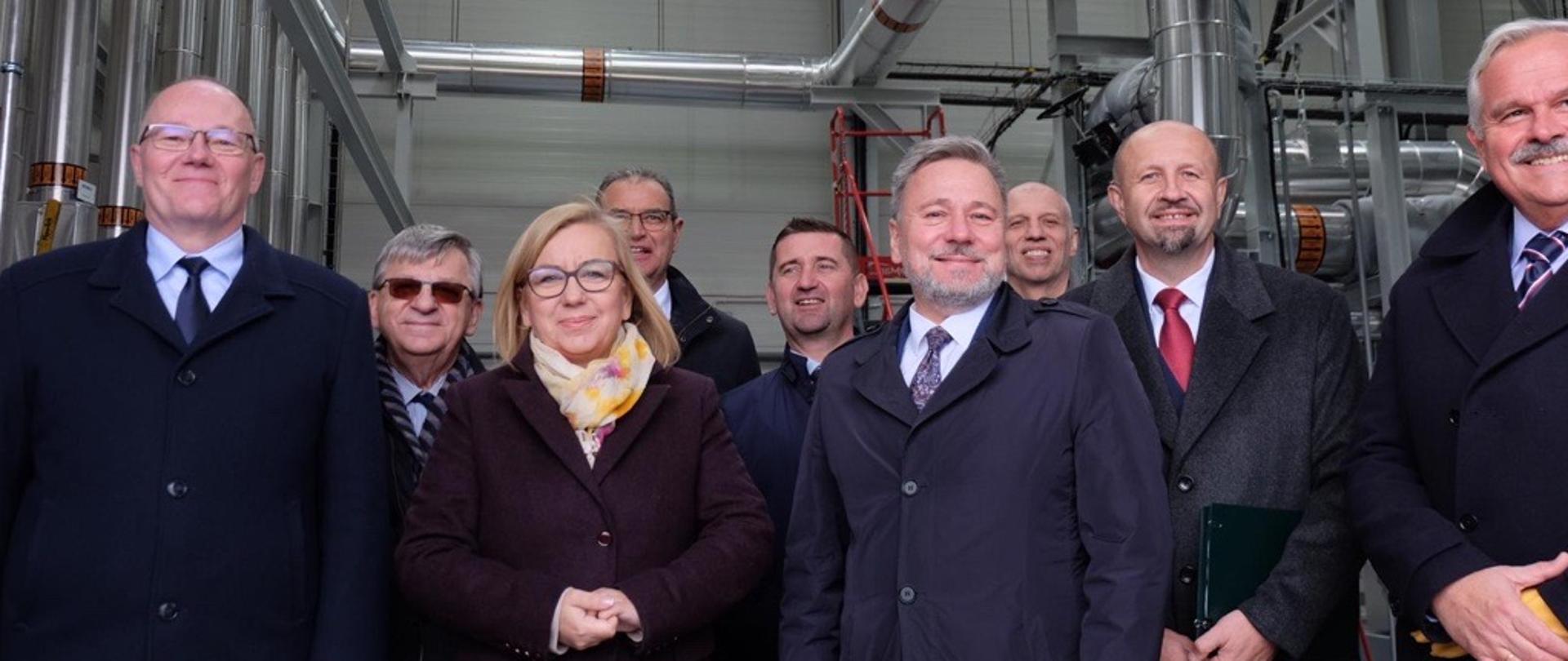 Na zdjęciu uczestnicy oficjalnego otwarcia ciepłowni geotermalnej w Koninie. Po lewej stronie Krzysztof Galos, Podsekretarz Stanu, Główny Geolog Kraju. Obok niego Paulina Hennig-Kloska, Minister Klimatu i Środowiska. Po prawej stronie, a także z tylu pozostali uczestnicy oficjalnego otwarcia geociepłowni. 