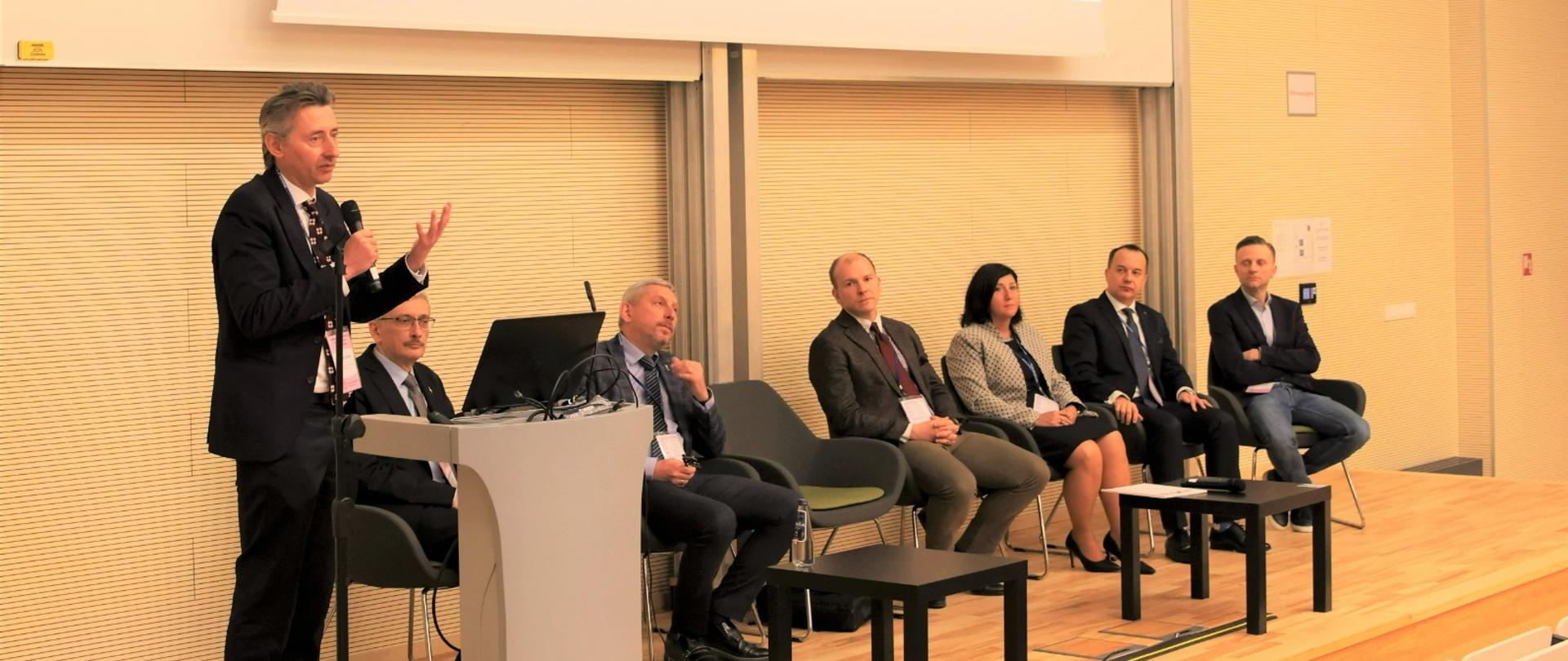 Widok z boku pod kątem, na podwyższeniu za mównica stoi wiceminister Gdula i mówi do mikrofonu, obok niego na krzesłach siedzi sześć osób.