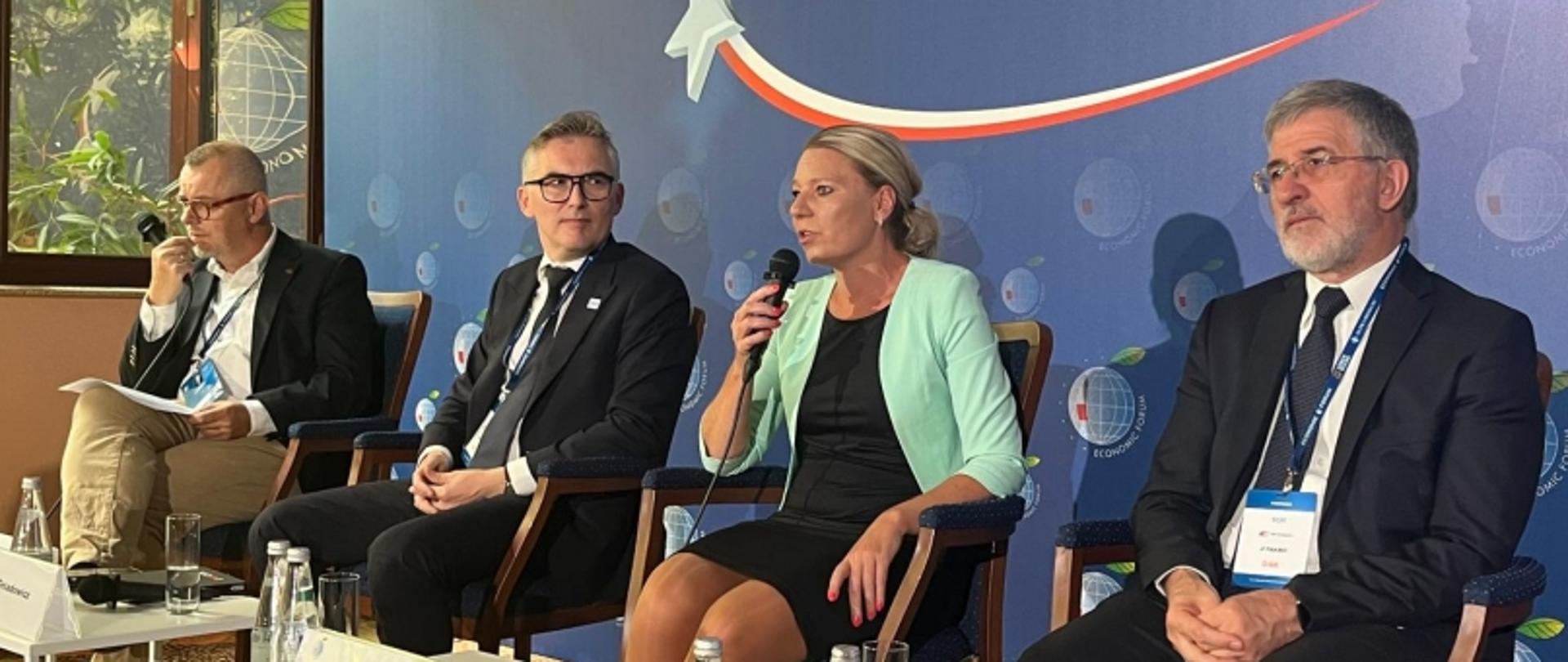 Panel konferencyjny z udziałem czterech osób, w tym Kamila Kamińskiego, na tle grafiki z samolotem i logotypami.