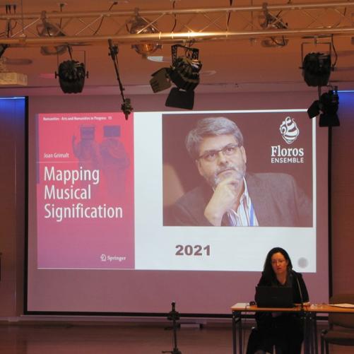 zdjęcie przedstawia prowadzącą wykład przemawiającą do mikrofonu przy biurku na scenie sali koncertowej, w tle ekran multimedialny