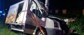 Zdjęcie przedstawia samochód dostawczy znajdujący się w przydrożnym rowie. Kabina kierowcy jest zdeformowana przez drzewo w które wjechał samochód. W tle widać ogrodzenie.