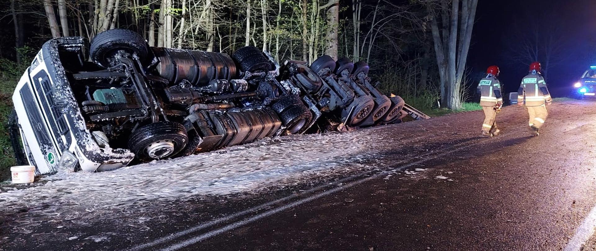 Zdjęcie przedstawia dwóch strażaków idących drogą DK 51 obok w przydrożnym rowie znajduję się uszkodzona cysterna znajdująca się w przydrożnym rowie.