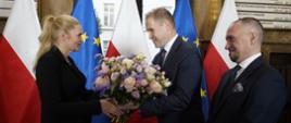 Minister Nowacka stoi naprzeciwko dwóch mężczyzn w garniturach, jeden z nich wręcza jej bukiet kwiatów, w tle polskie i unijne flagi.