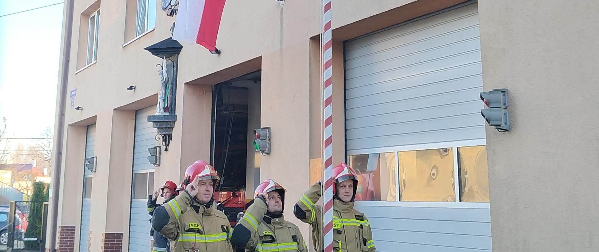 Przy budynku stoi kilku strażaków, przed nimi poczet flagowy salutuje przy maszcie z flagą Polski. Przed masztem zielone przycięte krzewy. Po lewej asfaltowy plac.

