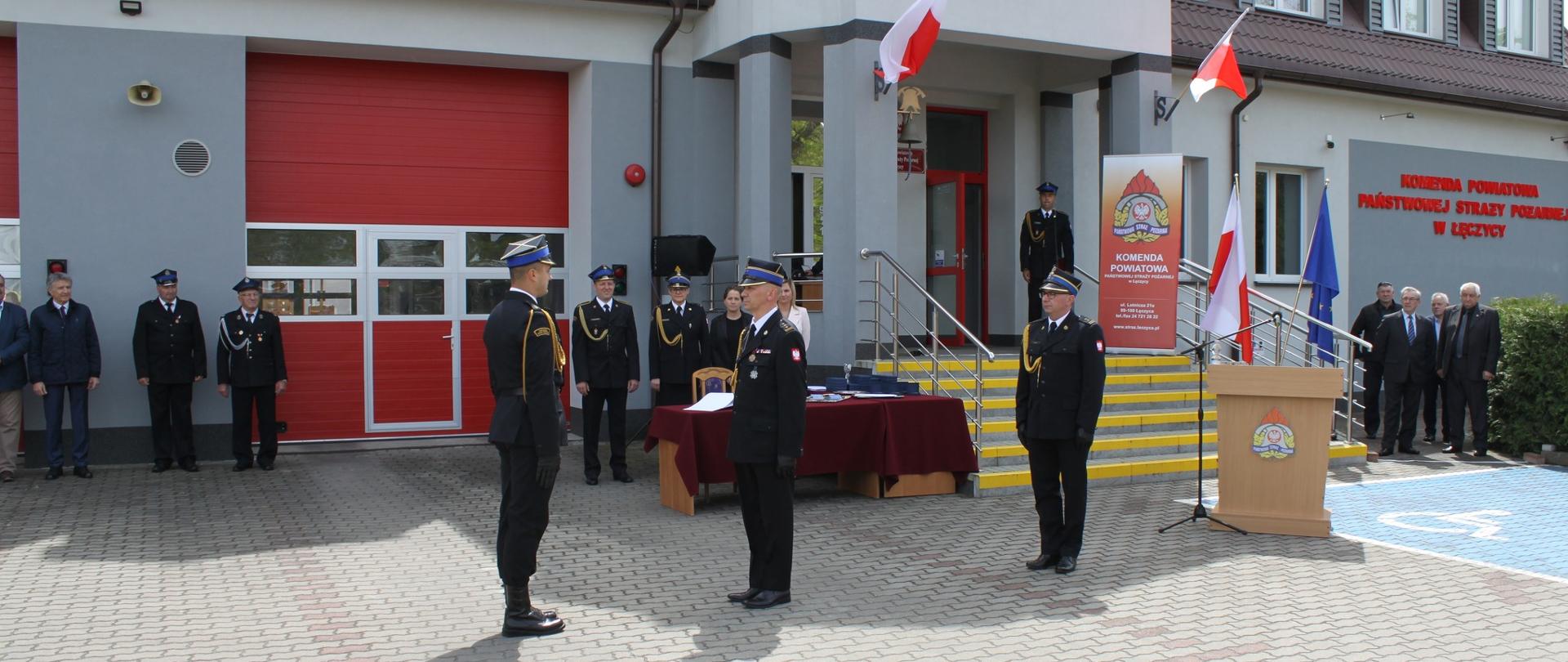 Na zdjęciu widać grupę strażaków oraz osób cywilnych stojących przed budynkiem komendy. Na budynku wywieszone są flagi Polski. Przed budynkiem na stojaku jest flaga Polski i Unii Europejskiej. Obok schodów stoi mównica z logo Państwowej Straży pożarnej, mikrofon oraz stół z czerwonym nakryciem. Jest widno.