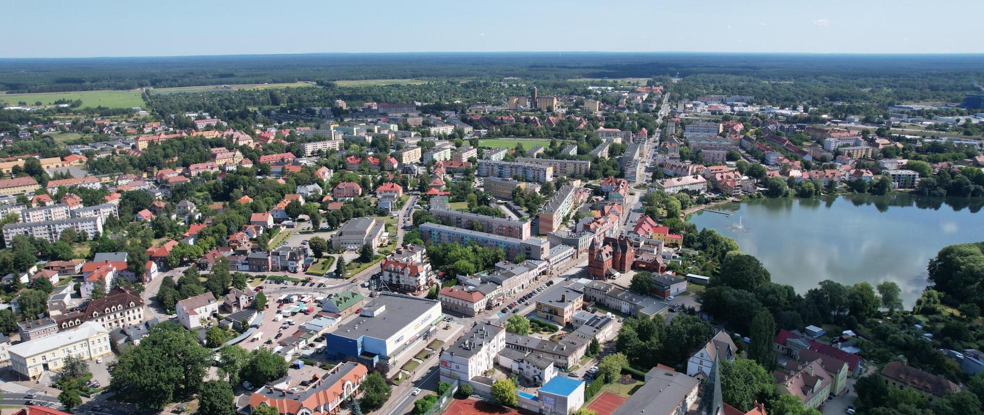 Widok z lotu ptaka na miasto. Widać różnorodne budynki, ulice oraz obszary zielone. Po prawej stronie znajduje się duże akwen wodny. Architektura obejmuje domy mieszkalne z czerwonymi dachami, większe budynki wielopiętrowe oraz ulice obsadzone drzewami. Okolica jest otoczona bujną zielenią i przestrzeniami otwartymi.