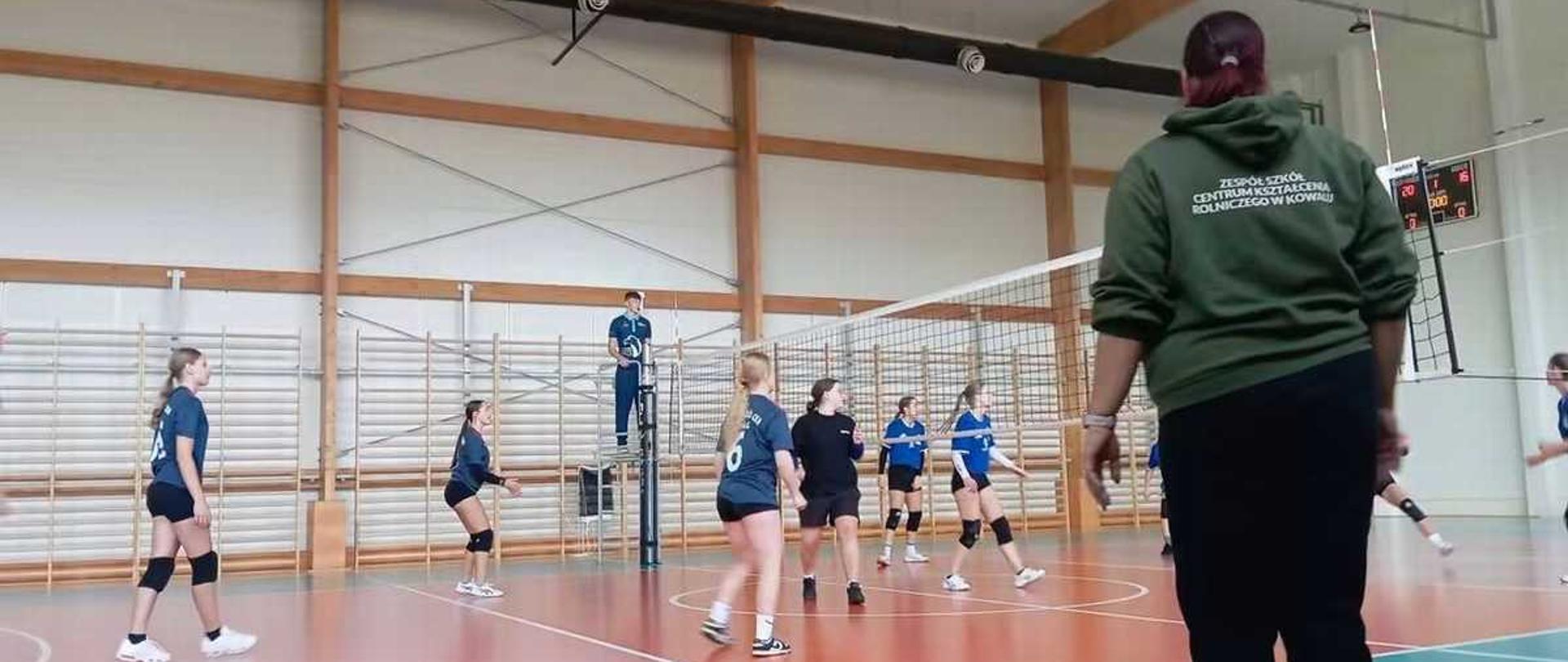 "Na pierwszym planie, po prawej stronie, tyłem do obiektywu stoi kobieta w zielonej bluzie z kapturem i czarnych spodniach, która obserwuje grę. Na plecach jej bluzy widnieje biały napis: „ZESPÓŁ SZKÓŁ CENTRUM KSZTAŁCENIA ROLNICZEGO W KOWALU”.
W głębi kadru, za siatką siatkarską, widać dwie drużyny zawodniczek w trakcie akcji.
• Dziewczęta po lewej stronie mają na sobie ciemnoniebieskie koszulki i czarne spodenki. Jedna z nich stoi przygotowana do odbioru po lewej stronie, druga dynamicznie porusza się w głębi boiska.
• Po prawej stronie siatki poruszają się zawodniczki w czarnych oraz jaśniejszych, niebieskich strojach.
• W tle, na podwyższeniu przy słupku sędziowskim, stoi sędzia w niebieskiej koszulce, uważnie śledzący przebieg gry.
Otoczenie i hala:
Gra toczy się na nowoczesnym boisku z dwukolorową nawierzchnią – pole gry jest koralowo-rude, natomiast otoczenie boiska ma kolor morski (zielono-niebieski). Linie wyznaczające pole gry są białe. Ściany hali są jasne, wykończone poziomymi, drewnianymi belkami konstrukcyjnymi. W tle po lewej stronie widać drabinki gimnastyczne, a po prawej stronie na ścianie wisi elektroniczna tablica wyników wyświetlająca czerwone cyfry. Pod wysokim sufitem przebiegają czarne rury instalacyjne oraz wiszą lampy oświetlające całą przestrzeń."