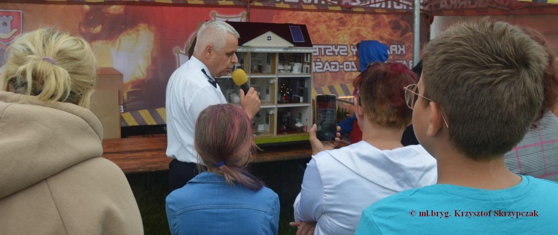 Edukacja z wykorzystaniem symulatora zagrożeń. Stadion sportowy w Miejskiej Górce, na którym odbywa się festyn rodzinny. Pod namiotem znajduje się symulator zagrożeń (dom jednorodzinny). Zastępca komendanta powiatowego PSP w Rawiczu prezentuje uczestnikom festynu zagrożenia, z jakimi mogą spotkać się przebywając w domu.