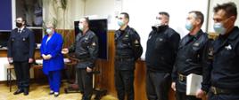 Widok z boku. W jednej linii obok siebie stoi sześciu strażaków PSP i OSP w umundurowaniu i maseczkach ochronnych oraz wicestarosta wadowicka w niebieskim ubraniu cywilnym. 