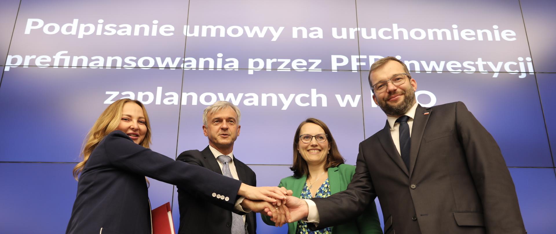 Cztery osoby, w tym minister Grzegorz Puda, stoją obok siebie przed stołem i trzymają się za ręce. W tle biały napis na niebieskim tle.