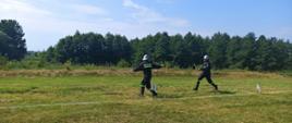 Zdjęcie przedstawia strażaków OSP biegnących podczas sztafety i podających sobie prądownice. Druhowie ubrani są w czarne stroje koszarowe, i biały hełm. Biegną po boisku z trawy.