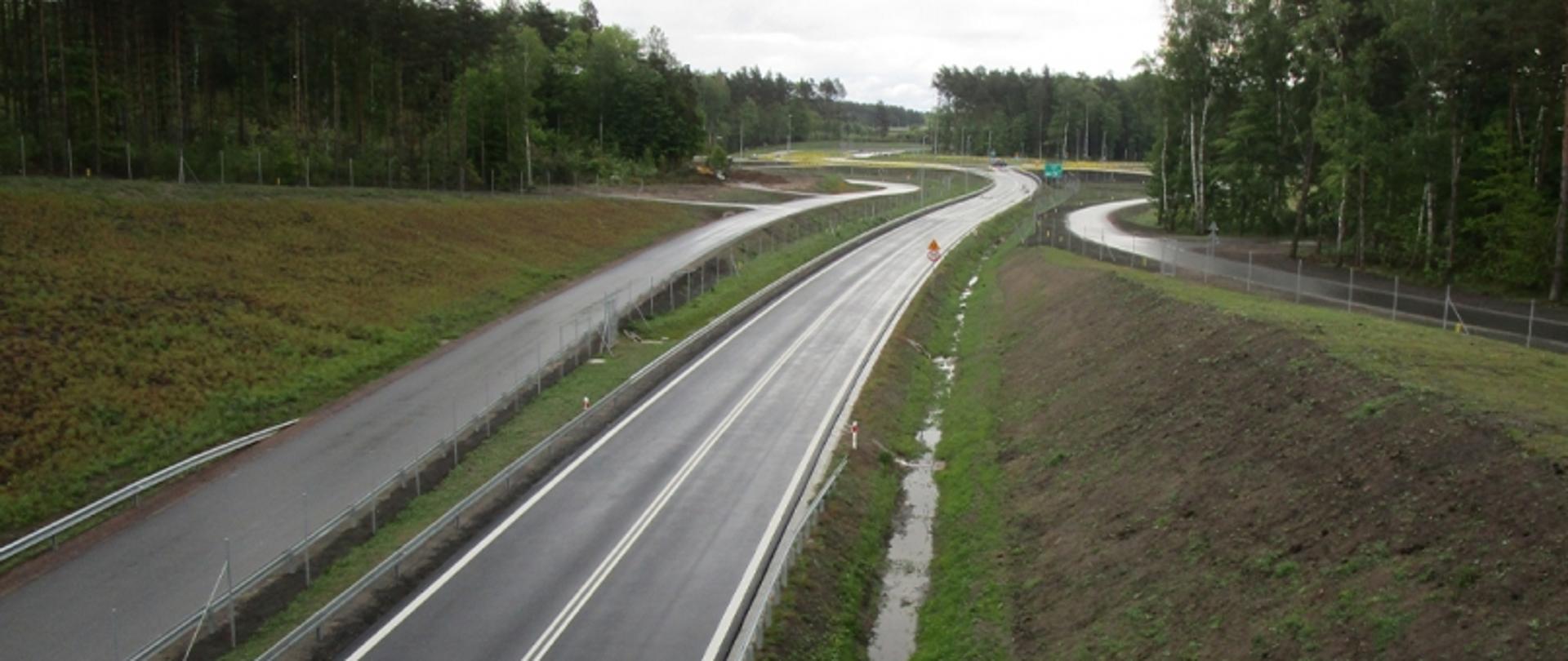 Nowo wybudowany odcinek drogi w Szczecinku otoczony lasem, dopuszczony do użytkowania przez nadzór budowlany. Droga biegnie przez pagórkowaty teren z zabezpieczeniami i odwodnieniem.