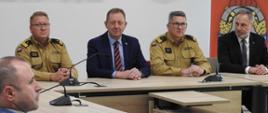 Dwóch strażaków i dwóch mężczyzn w garniturach siedzi przy stole. Spoglądają na mężczyznę siedzącego po ich prawej stronie. Za nimi jasna ściana. Na ścianie ciemne litery tworzące napis wiat czyński, herb powiatu opoczyńskiego oraz czerwony plakat z logo PSP. Na stołach ustawione są mikrofony. 