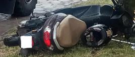Wypadek motorowerzysty. Rawicz, ul. Armii Krajowej. Na trawniku obok drzewa leży motorower biorący udział w kolizji. Jest koloru czarnego. obok siedzenia znajduje się kask. Z lewej strony droga i fragment wozu strażackiego.