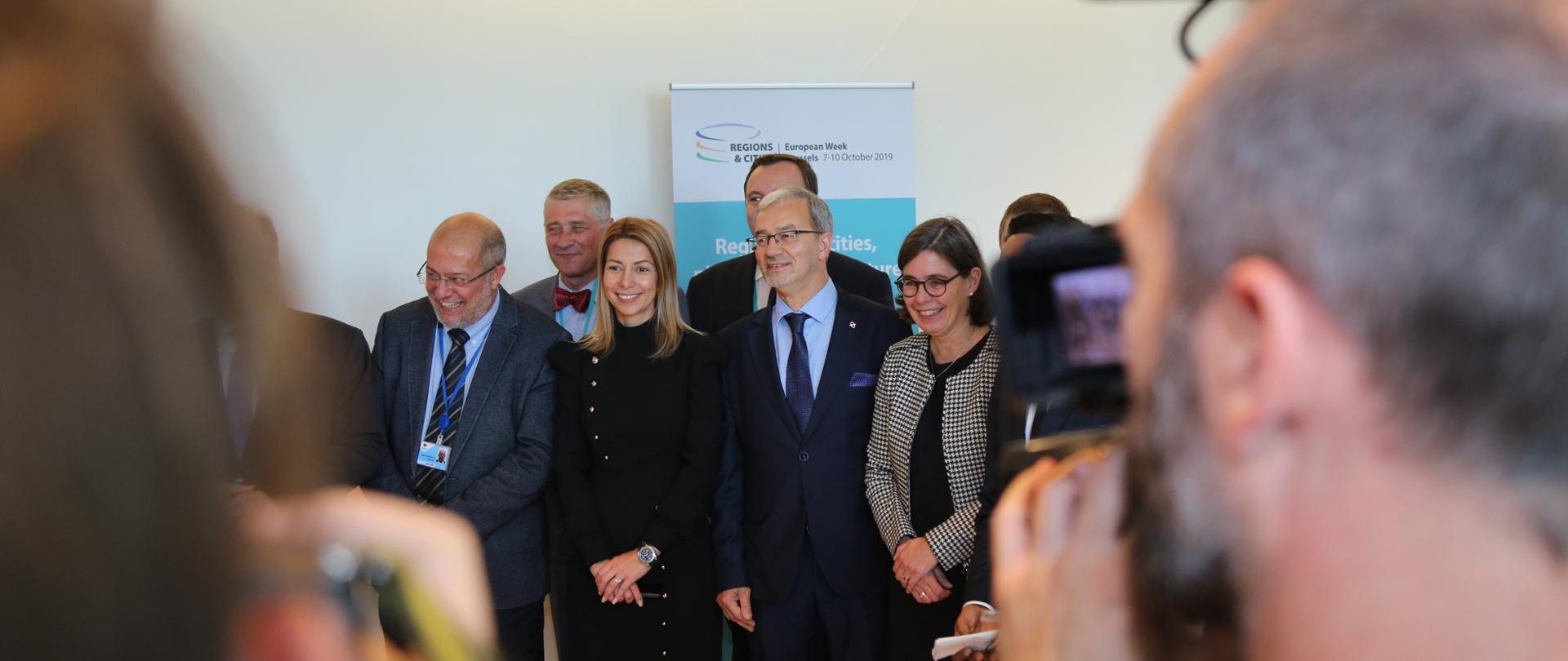 Minister Jerzy Kwieciński wraz z innymi uczestnikami spotkania stoi przed roll-upem, na którym napisane jest European Week of Regions and Cities. Przed ministrem stoją dziennikarze. 
