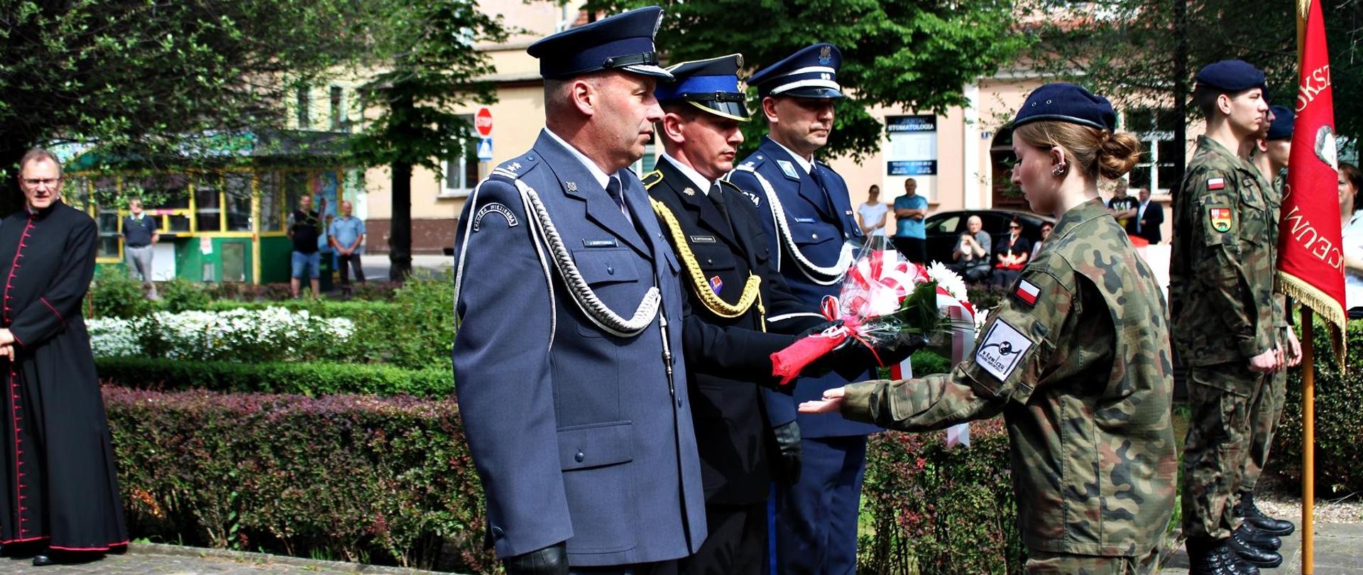 Święto Konstytucji Plac Wolności w Rawiczu. Na pierwszym planie delegacja Policji, PSP i SW z wiązanką kwiatów. Strażak przekazuje ją uczennicy klasy mundurowej, tzw. kwiaciarce. W tle budyki i drzewa.