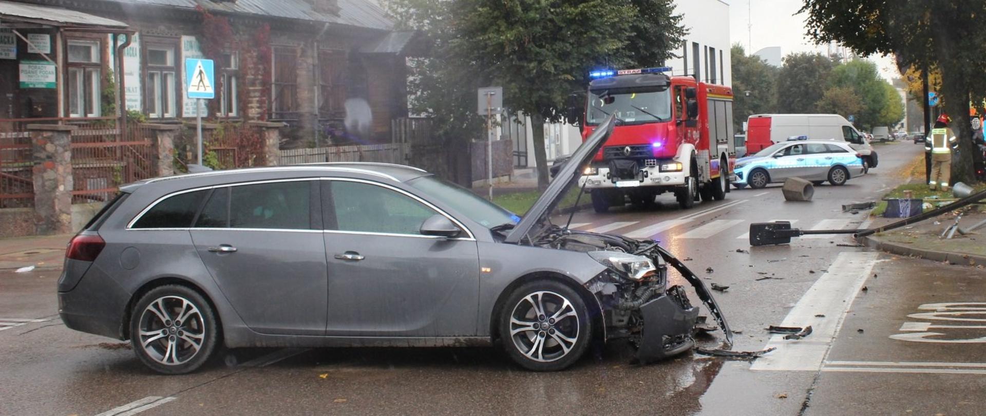 Samochód osobowy marki Audi stojący na środku skrzyżowania po zderzeniu z inny pojazdem. Samochód ma uszkodzony przód. W tle samochód ratowniczo-gaśniczy. Na jezdni leży wywrócony betonowy kosz na śmieci oraz złamana latarnia.
