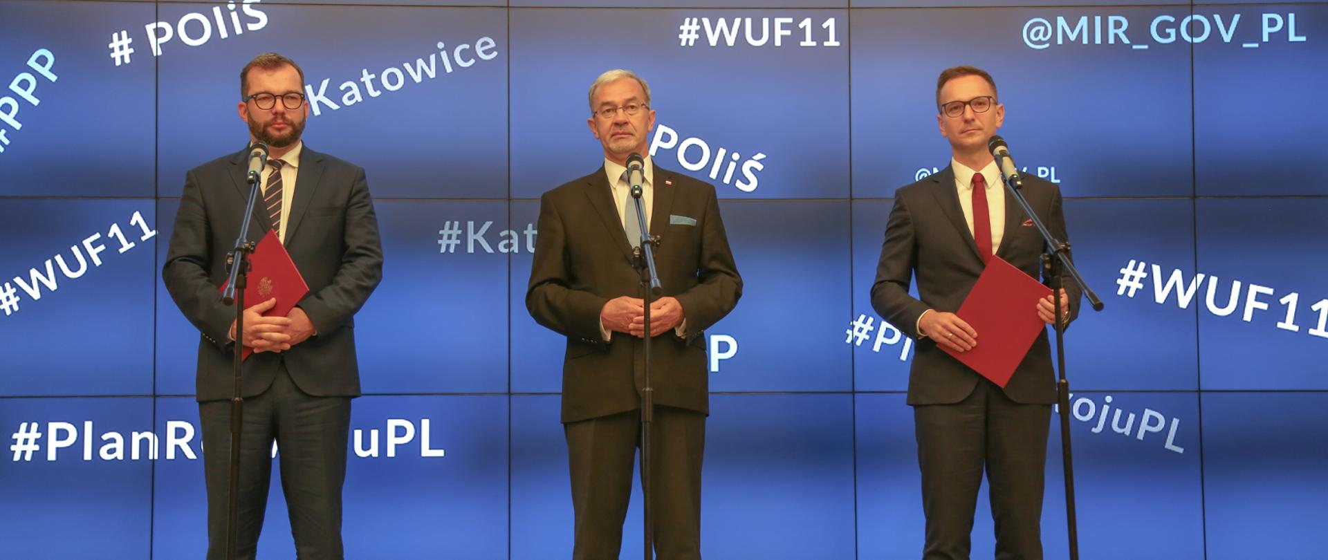 W sali konferencyjnej na scenie przed mikrofonami stoją: minister inwestycji i rozwoju Jerzy Kwieciński oraz wiceministrowie Waldemar Buda i Grzegorz Puda. Za nimi niebieska ścianka.
